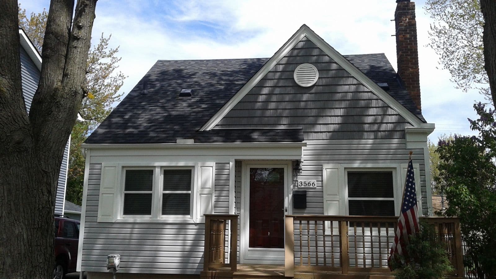 Charming gray house with front porch, American flag, and leafing trees in the background.