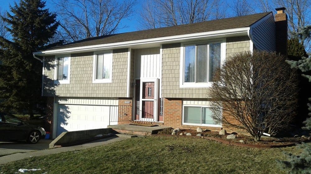 Split-level home with snow-covered yard and trees, featuring beige siding and dark green shutters.