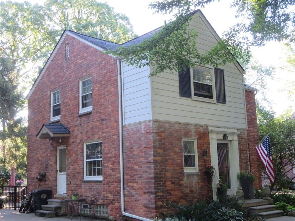 Charming brick and siding house with American flag, surrounded by greenery.