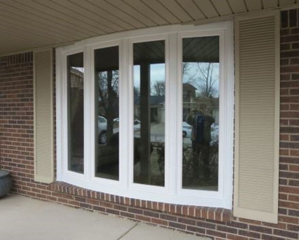Modern bay window with three large glass panels and beige shutters on a brick exterior.