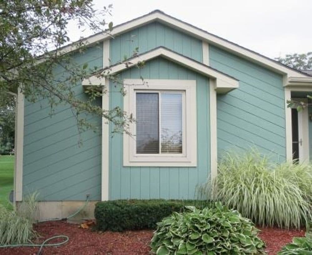 Teal house exterior with a modern window, surrounded by green bushes and mulch.