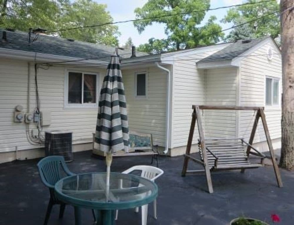 Backyard patio with a swing, table, chairs, and striped umbrella beside a house.