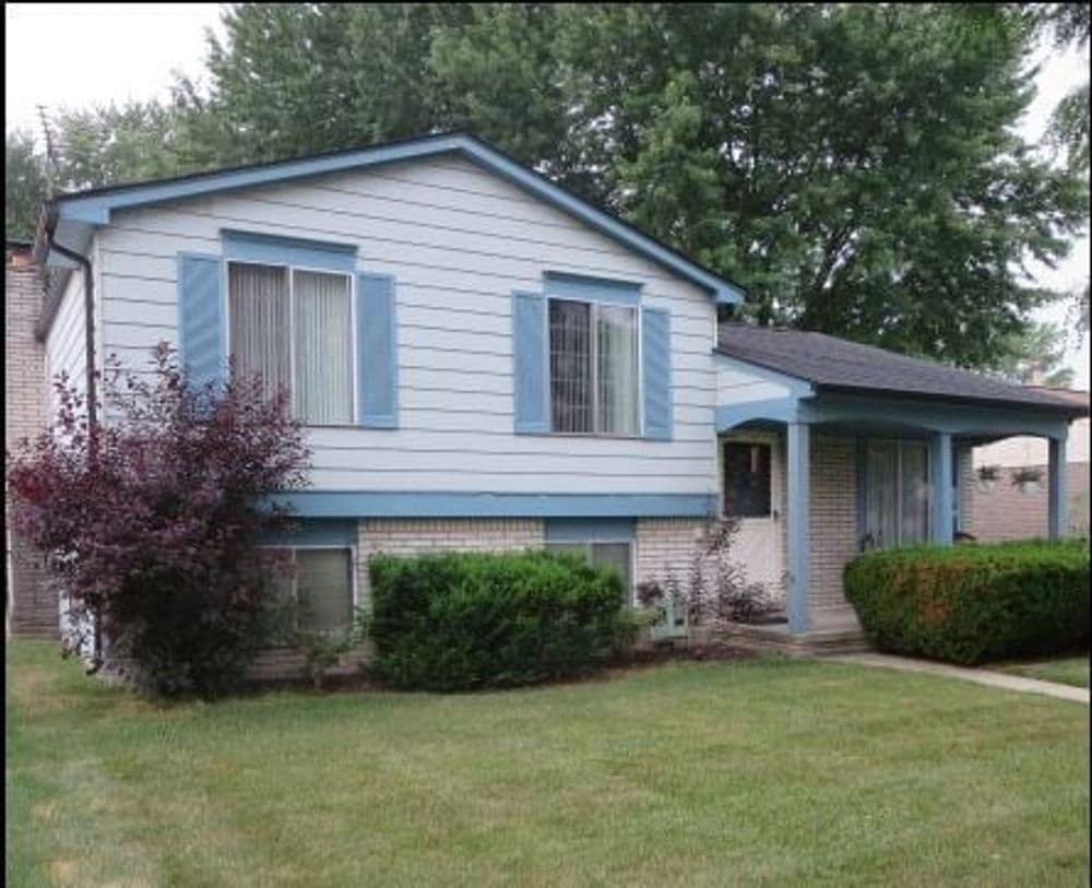 Two-story suburban house with blue trim, landscaped yard, and trees in the background.